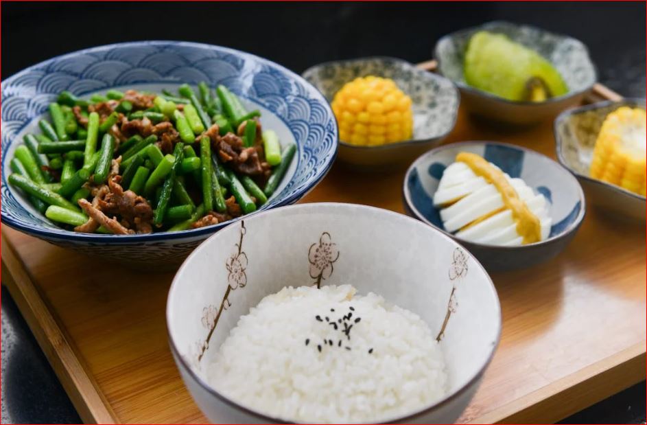 A bowl of white rice with a spoon and a green plant in the background